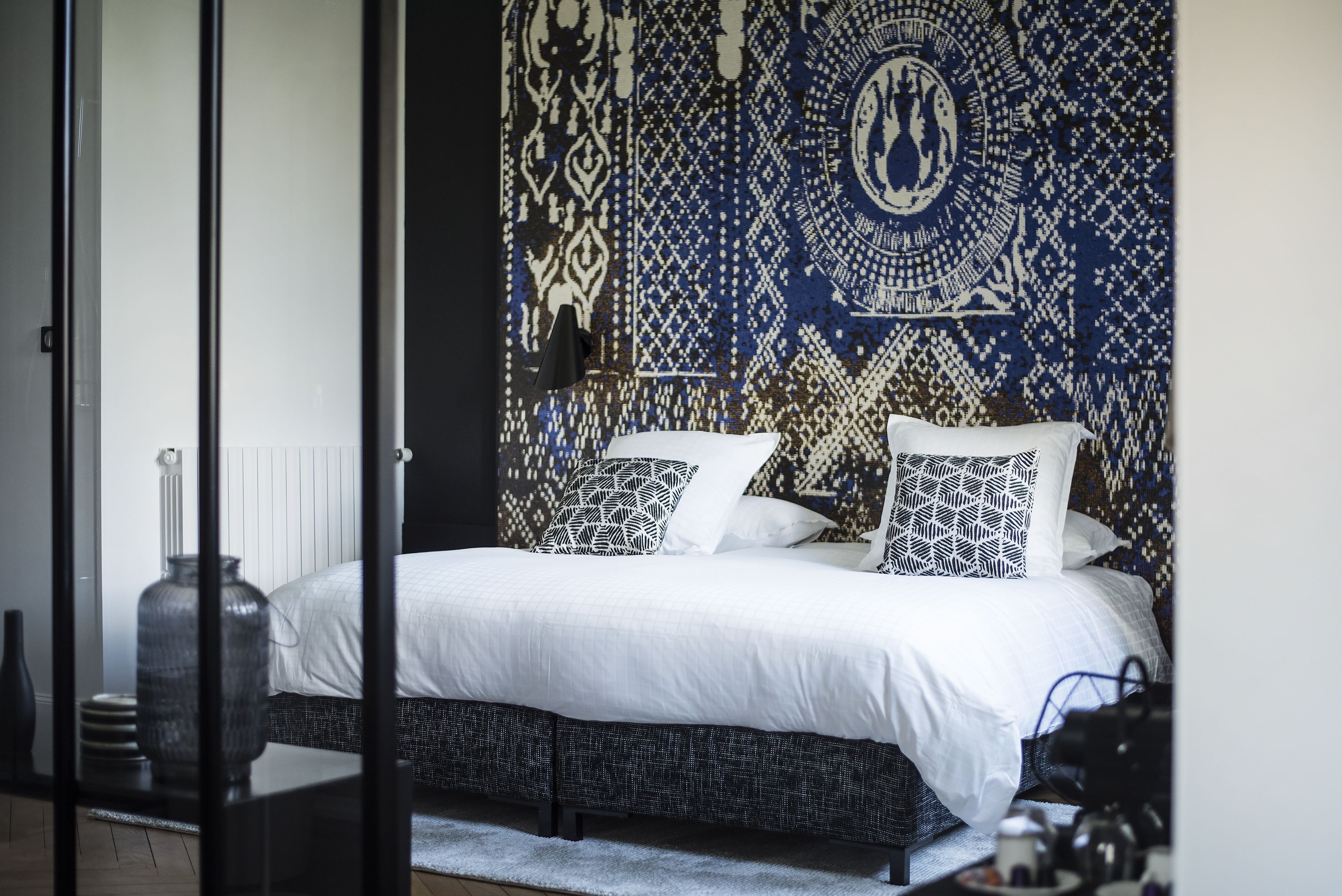 Une chambre à coucher avec une tapisserie murale à motifs, un lit avec une literie blanche et une lampe noire sur une table de chevet.