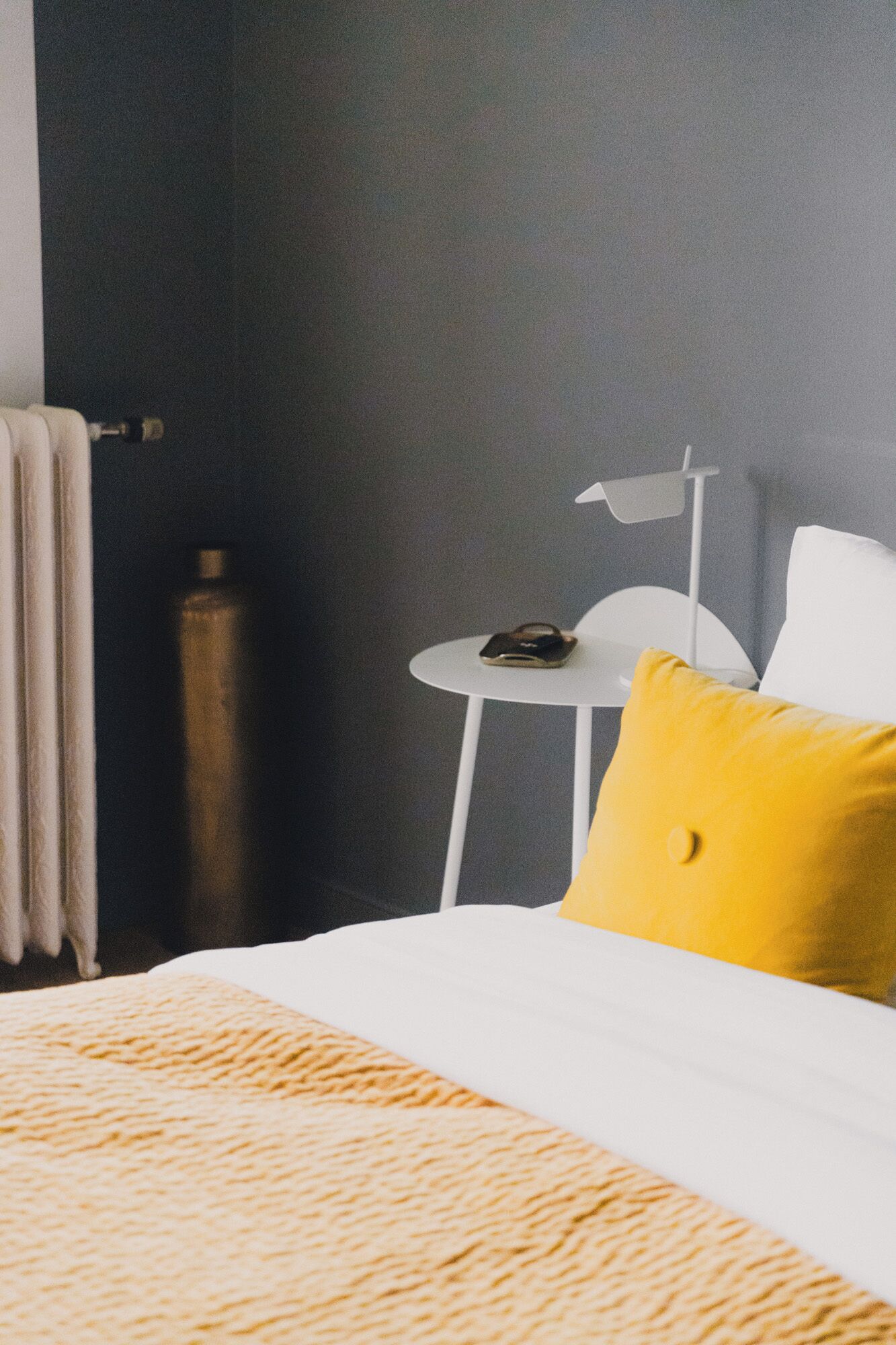 Une chambre d’hôtel avec un lit, une petite table blanche, un oreiller jaune, une lampe blanche et une serviette accrochée à un crochet.