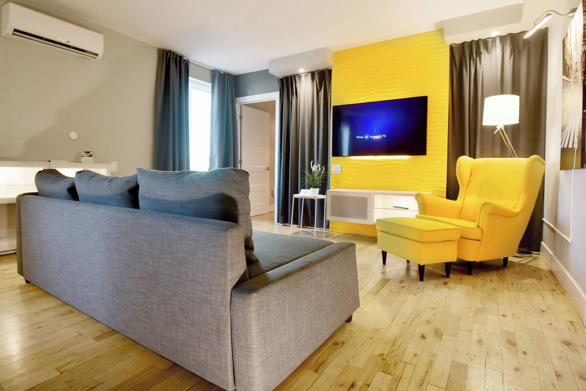 A modern living room with a grey sofa, a yellow armchair, a flat-screen TV mounted on a yellow accent wall, and a wooden floor.