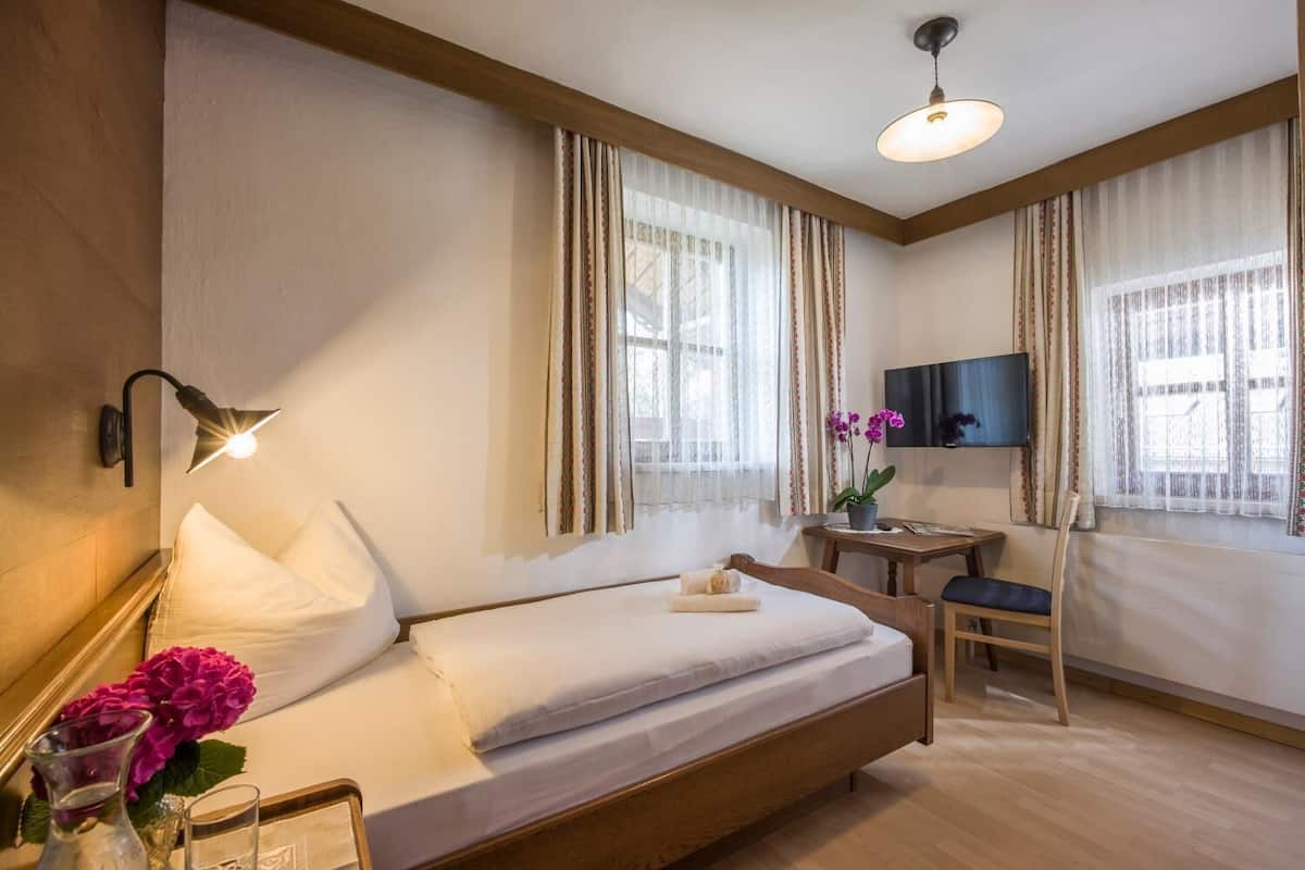 A hotel room with a bed, bedside lamp, TV, desk, chair, and a window with curtains.