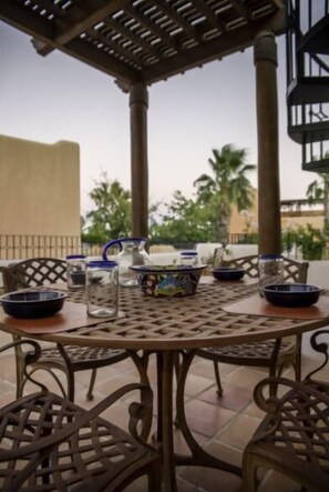 A round metal table with chairs, a pitcher, bowls, and a glass on a patio with a pergola overhead.