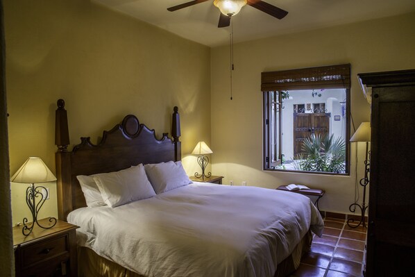 A bedroom with a wooden bed, two bedside tables with lamps, a ceiling fan, and a window with a view of the outside.