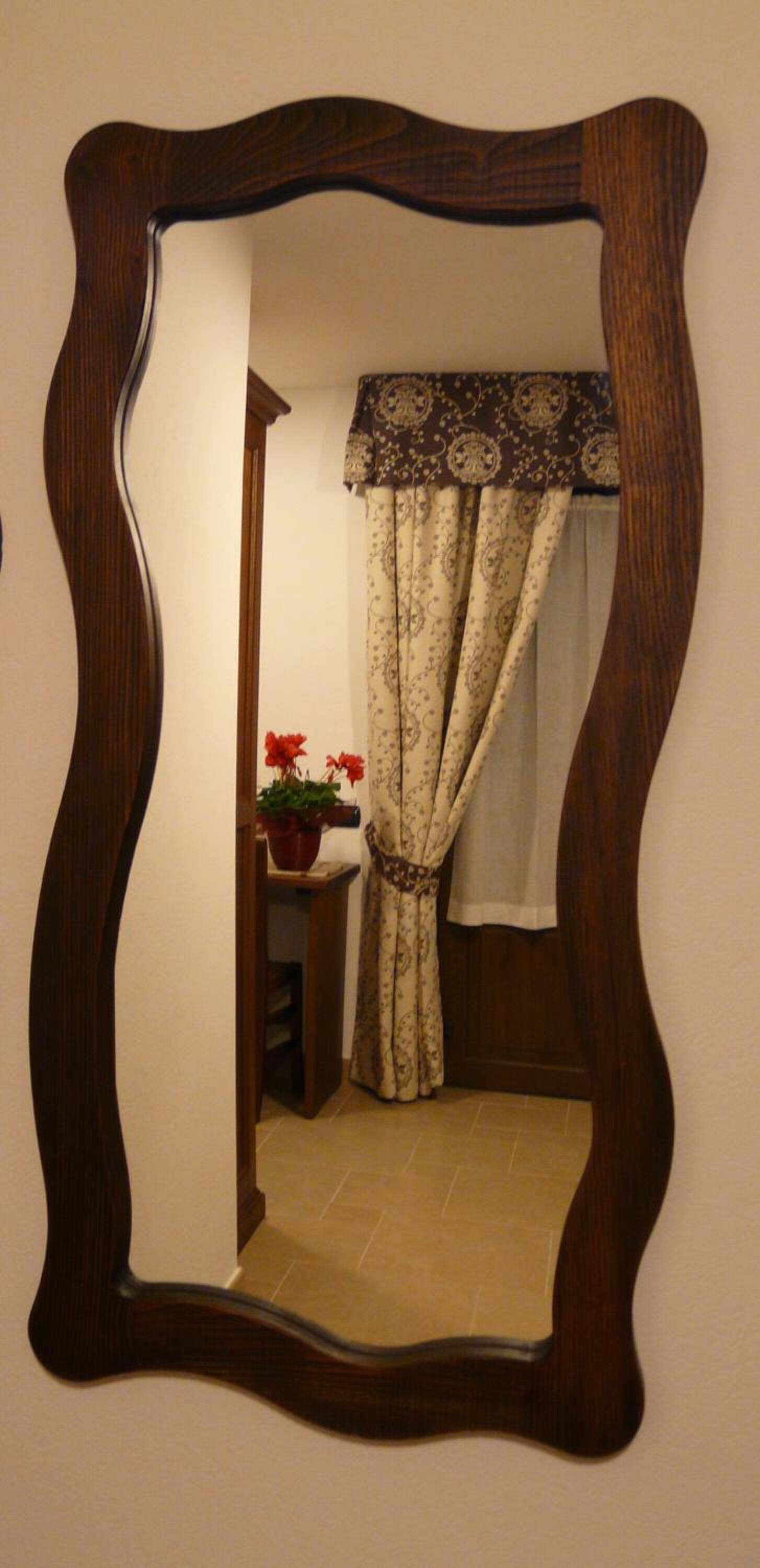 A mirror with a wavy frame reflecting a bathroom with a patterned curtain, a potted plant, and a small table.