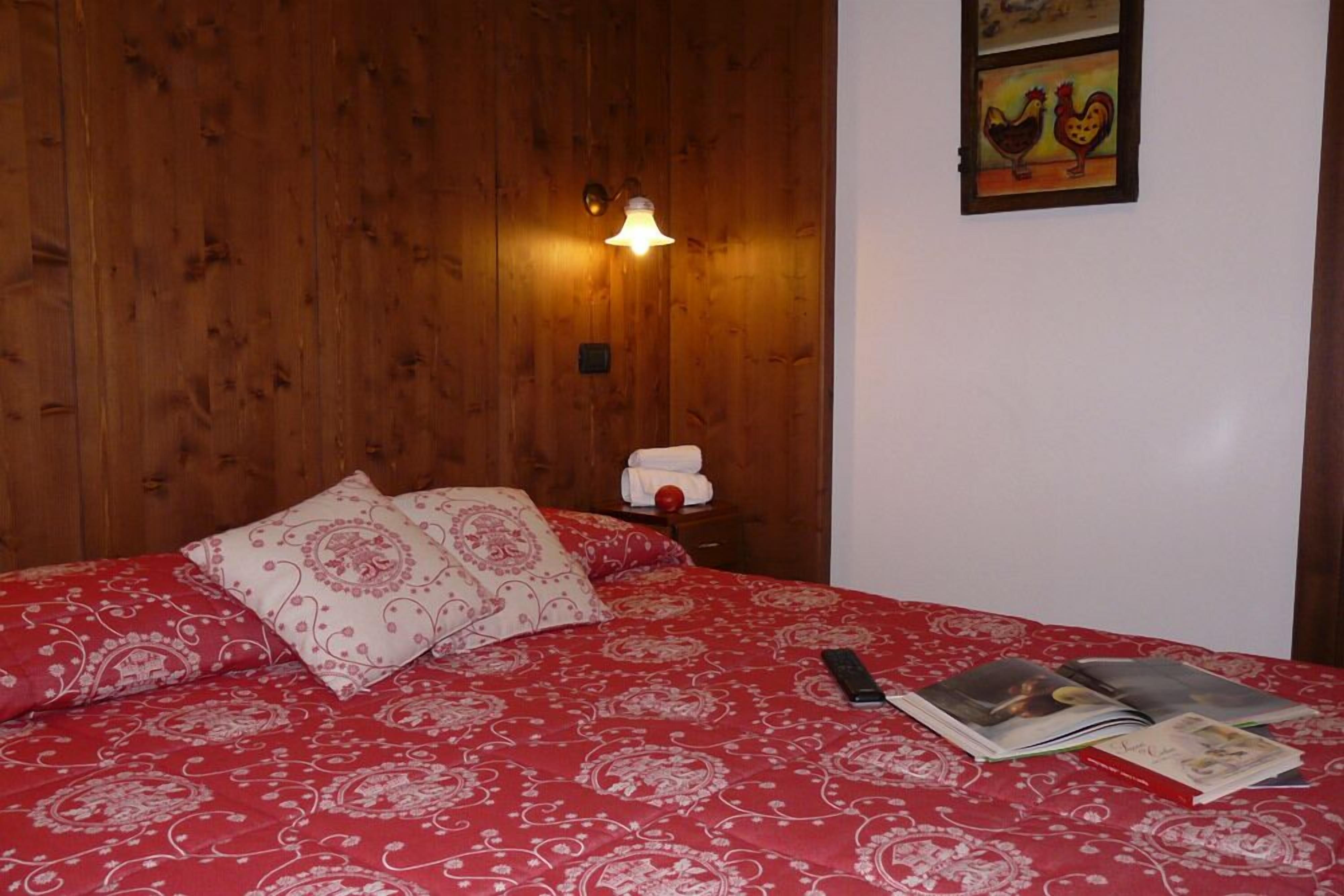 Une chambre à coucher avec une tête de lit en bois, une couvre-lit rouge à motifs, une lampe de chevet, une table de chevet avec une serviette et un objet rouge, et un mur orné d’un tableau encadré.