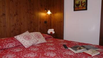 Une chambre à coucher avec une tête de lit en bois, une couvre-lit rouge à motifs, une lampe de chevet, une table de chevet avec une serviette et un objet rouge, et un mur orné d’un tableau encadré.