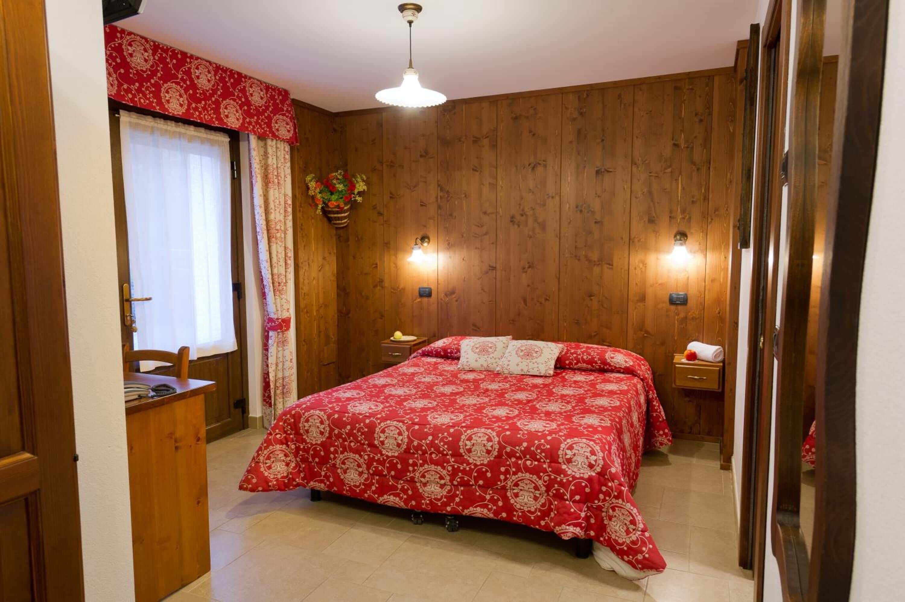 Une chambre avec un mur en bois, un lit recouvert d’une couvre-lit à motifs rouges et blancs, une table de chevet et une lampe suspendue.