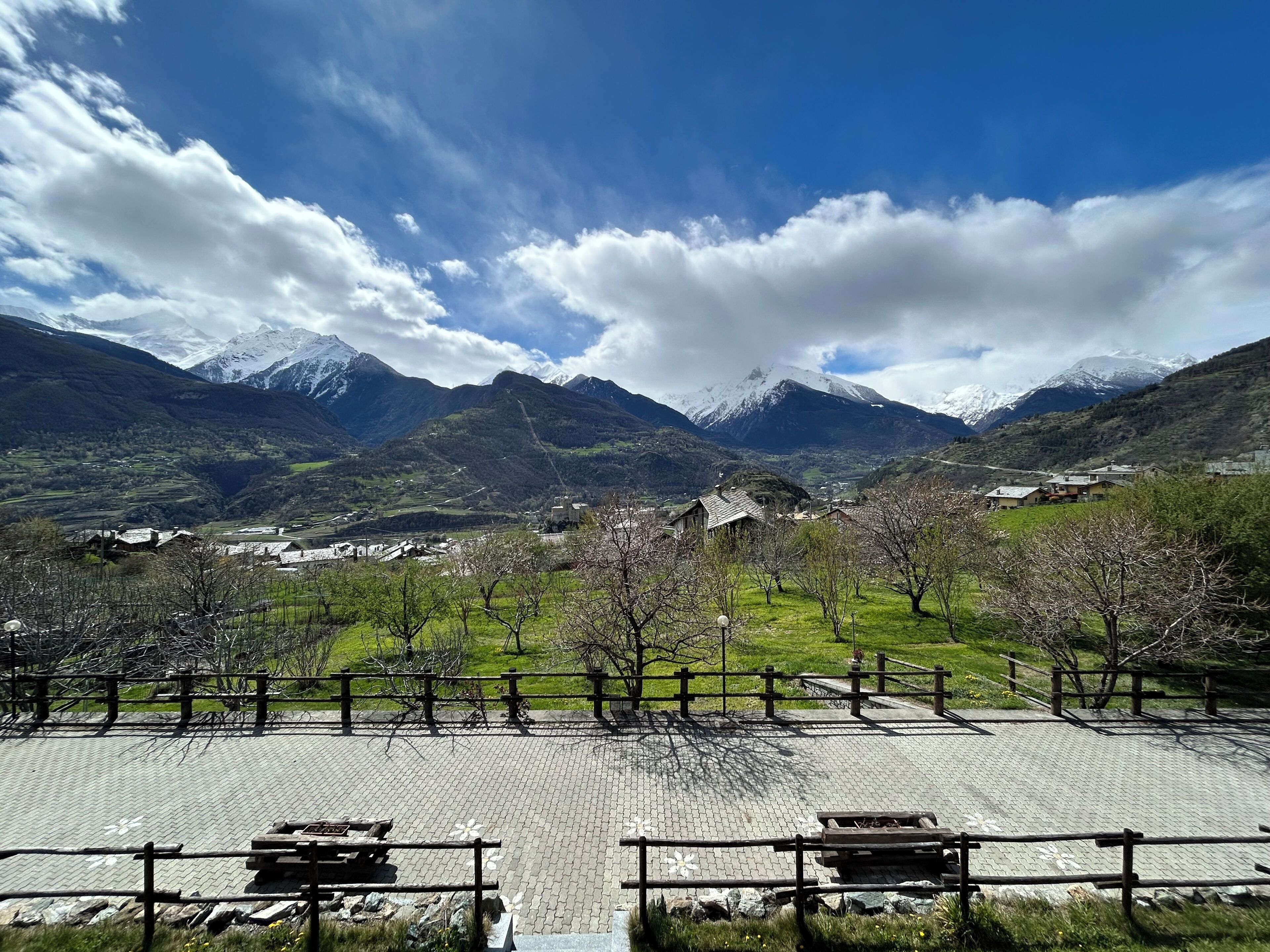 Un área pavimentada con bancos y una cerca, rodeada de césped verde y árboles, con un paisaje montañoso al fondo.
