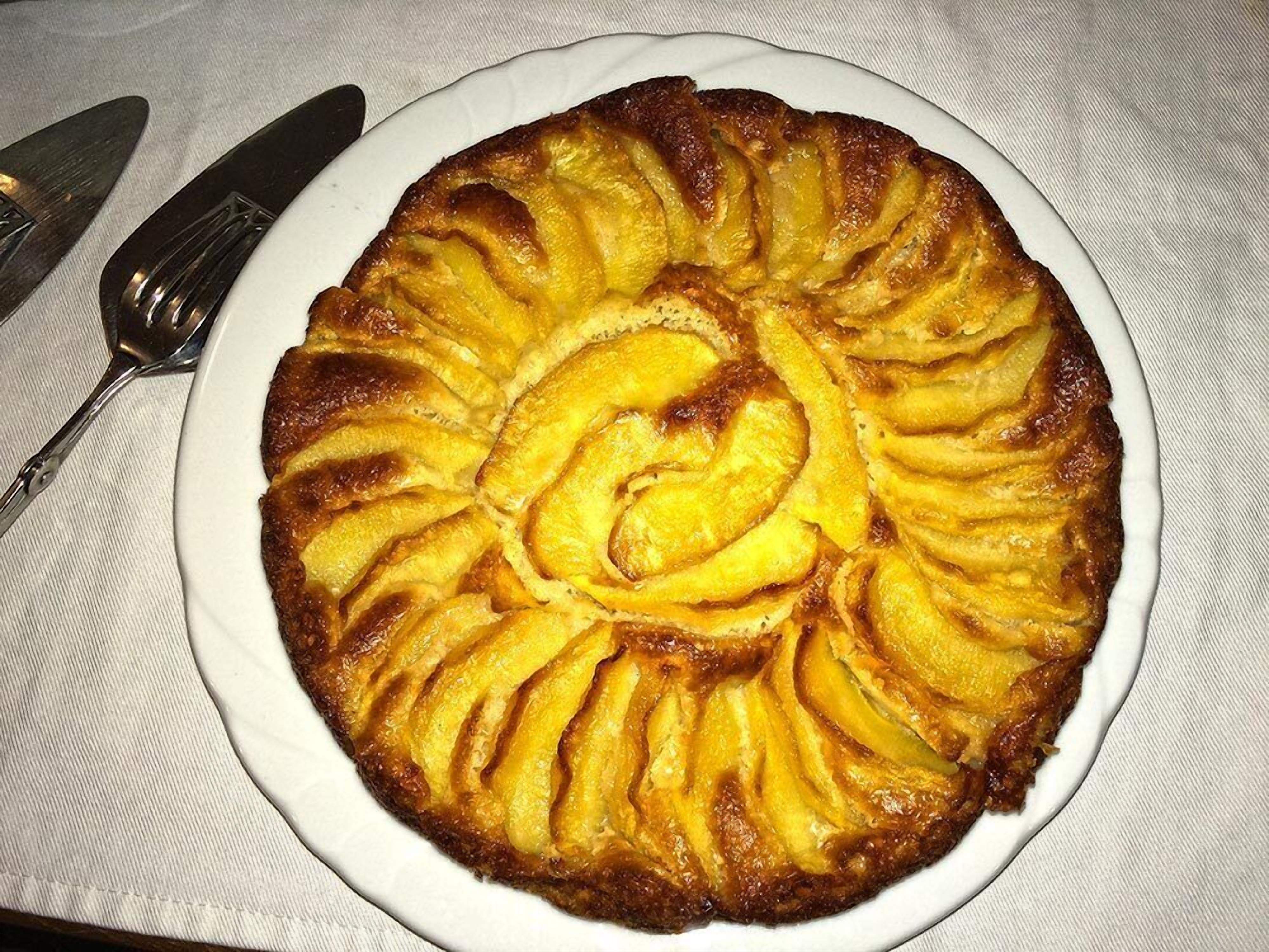 Ein runder Obstkuchen mit spiralförmlich angeordneten Fruchtscheiben auf einem weißen Teller.