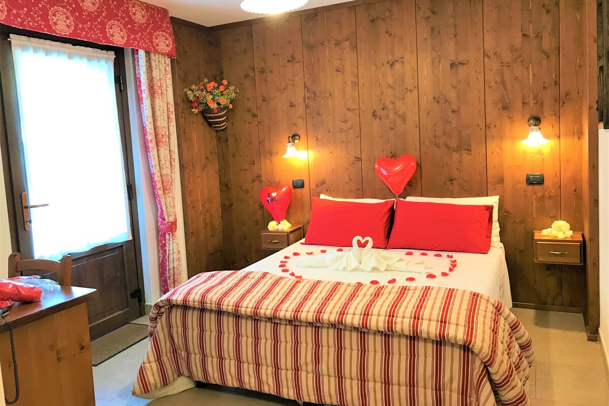 A cozy bedroom with a wooden wall, a striped bedspread, red pillows, heart-shaped balloons, and a bouquet of flowers.