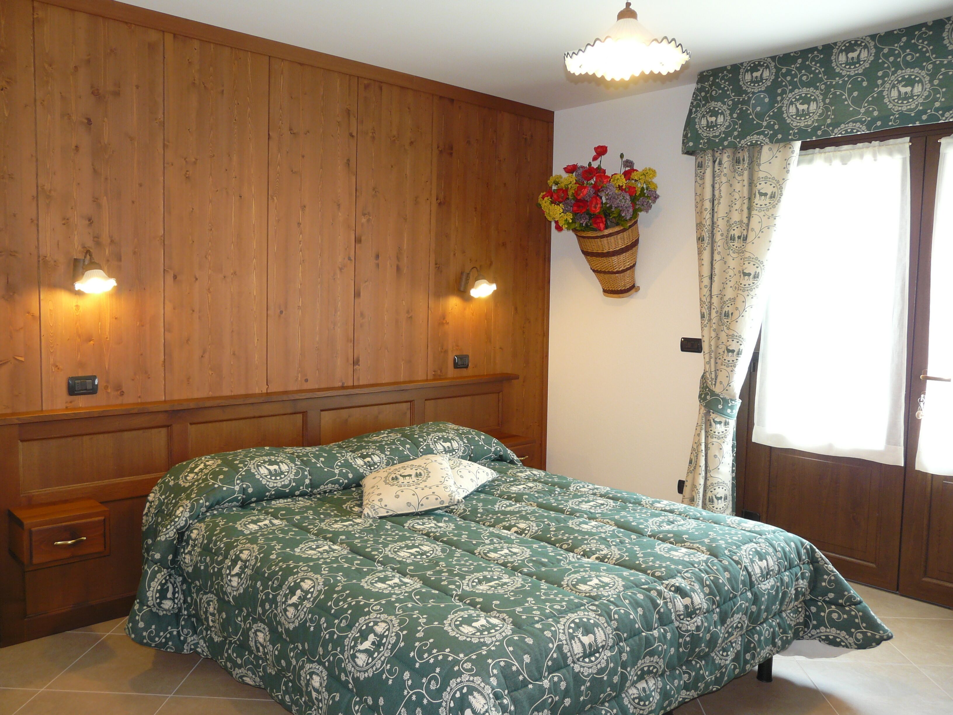 A bedroom with a wooden headboard, a green patterned bedspread, a bedside table, and a wall-mounted basket with flowers.
