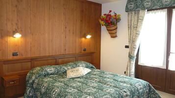 A bedroom with a wooden headboard, a green patterned bedspread, a bedside table, and a wall-mounted basket with flowers.