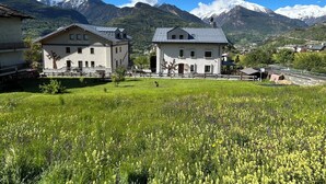 A rural setting with two buildings, one with a balcony and another with a sloped roof, surrounded by a field of yellow flowers and mountains in the background. - B&B Verger Plein Soleil (Saint-Pierre)