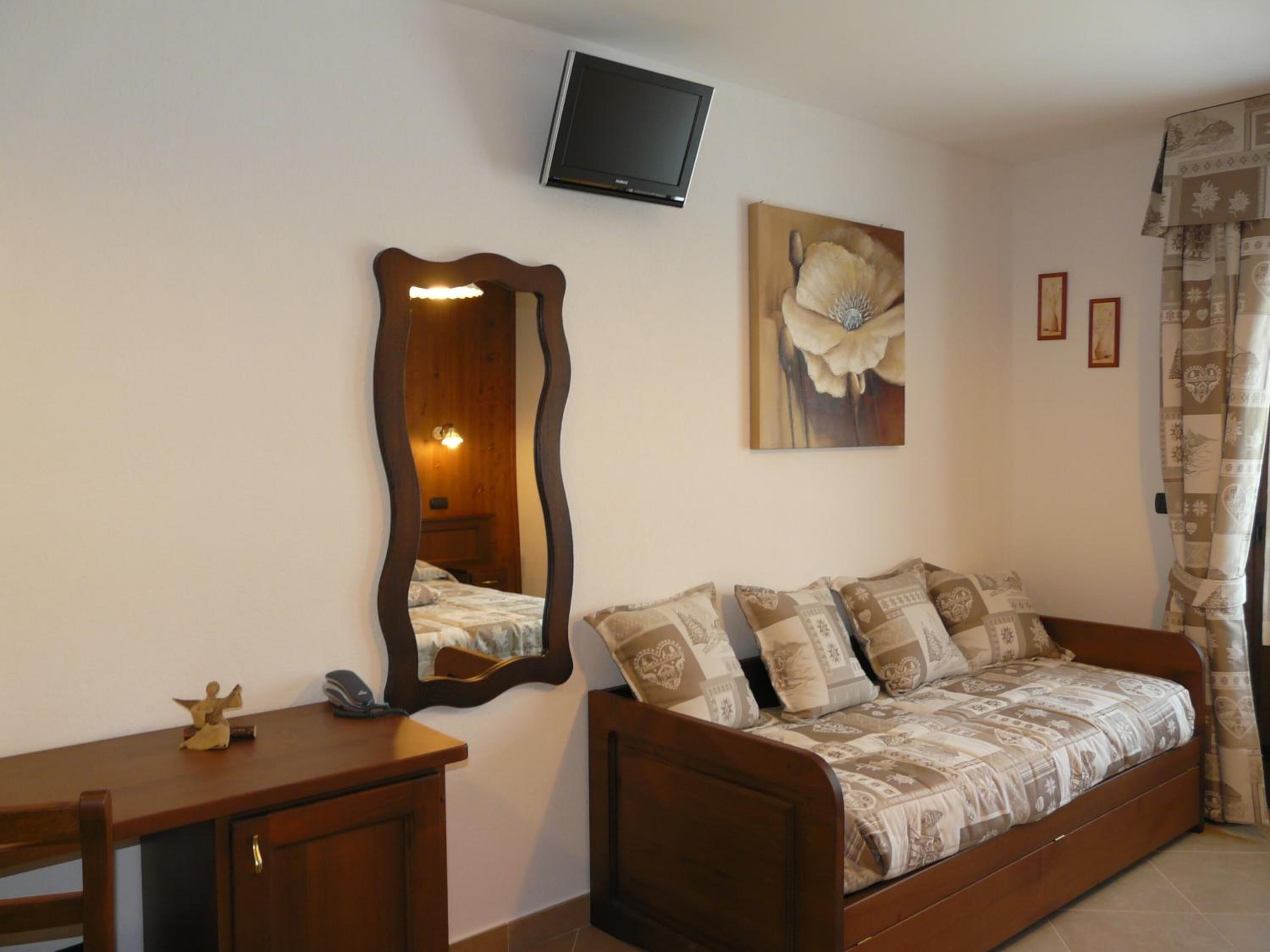 Ein Schlafzimmer mit einem Bett, einer Holzkommode, einem Spiegel, einem an der Wand befestigten Fernseher und einem Blumenbild.