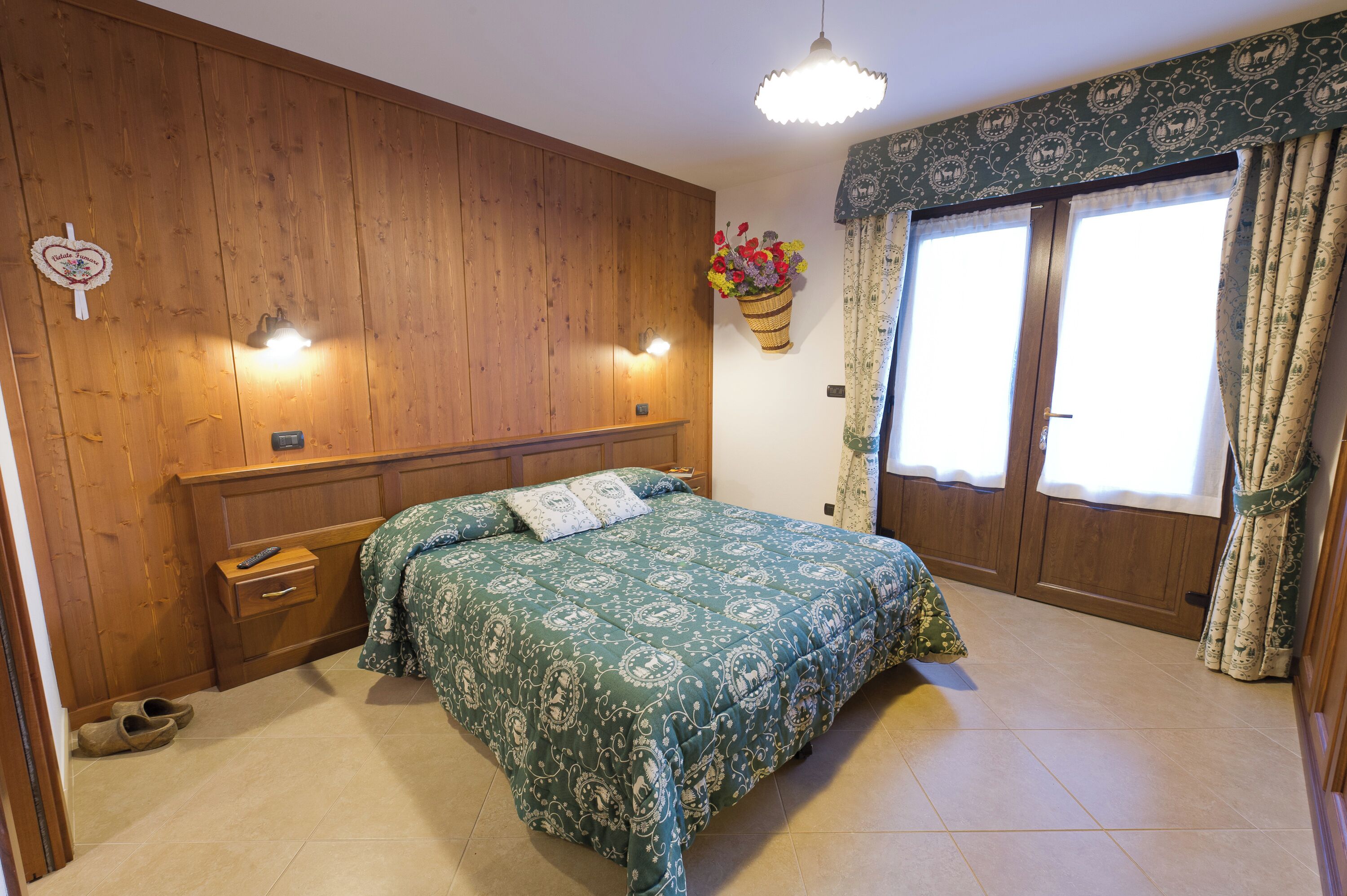 Un dormitorio con cabecera de madera, cama con colcha estampada, mesita de noche con lámpara y ventana con cortinas.