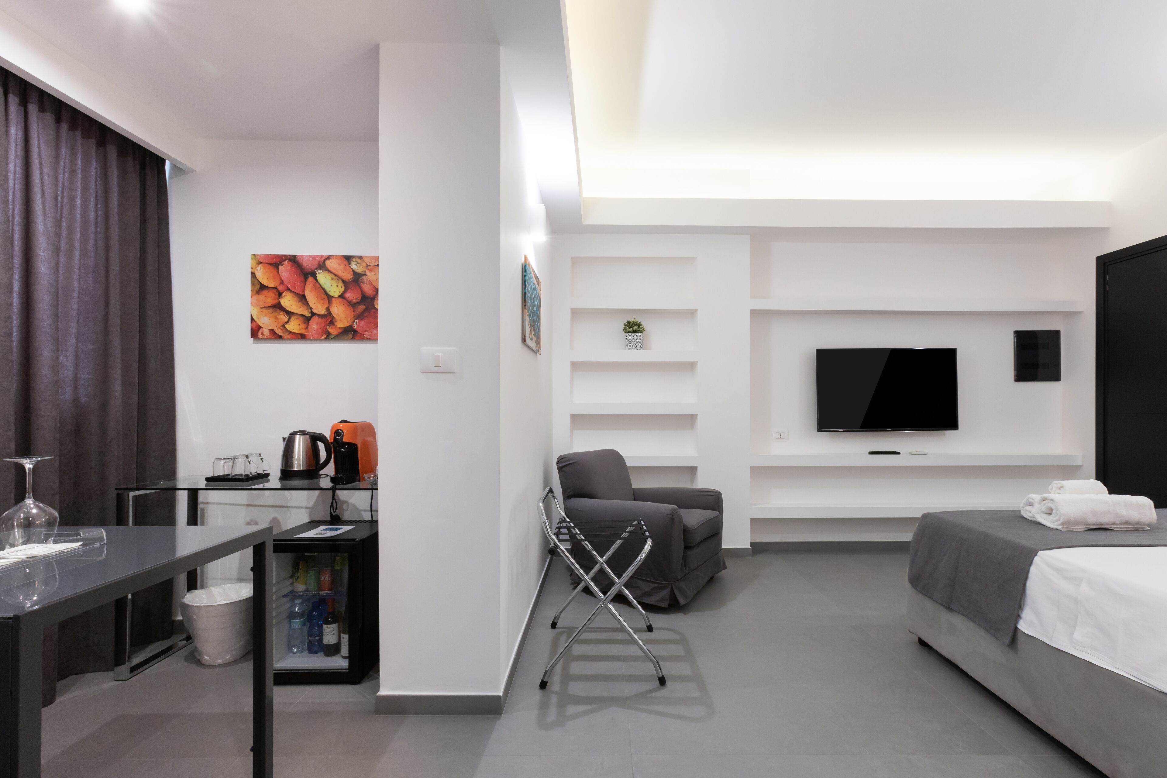 Een moderne hotelkamer met een bed, een televisie, een bank, een bureau met een waterkoker, en een klein plankje met een plant.