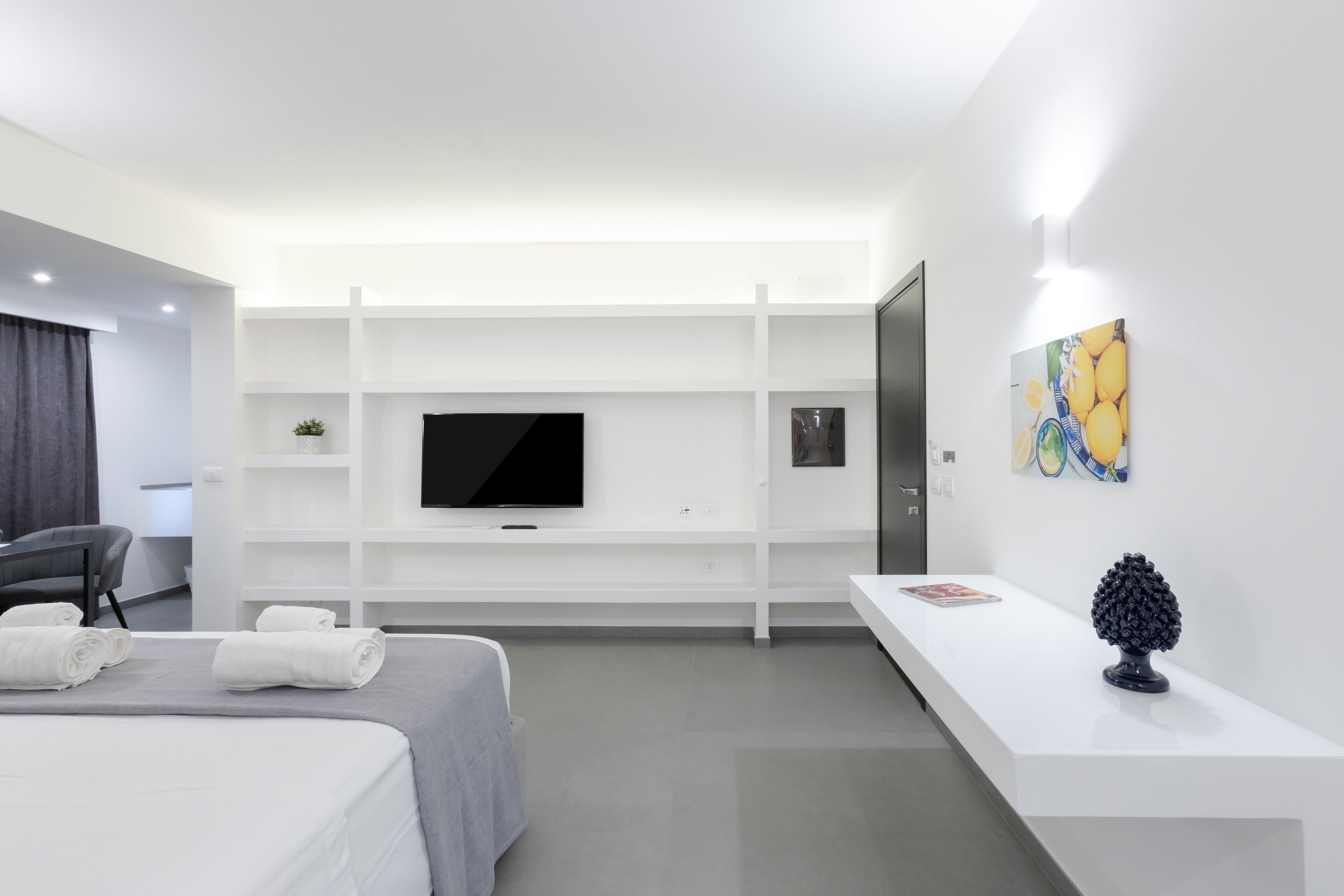 A modern hotel room with a bed, a TV mounted on a white wall, a desk with a lamp, and a small plant.