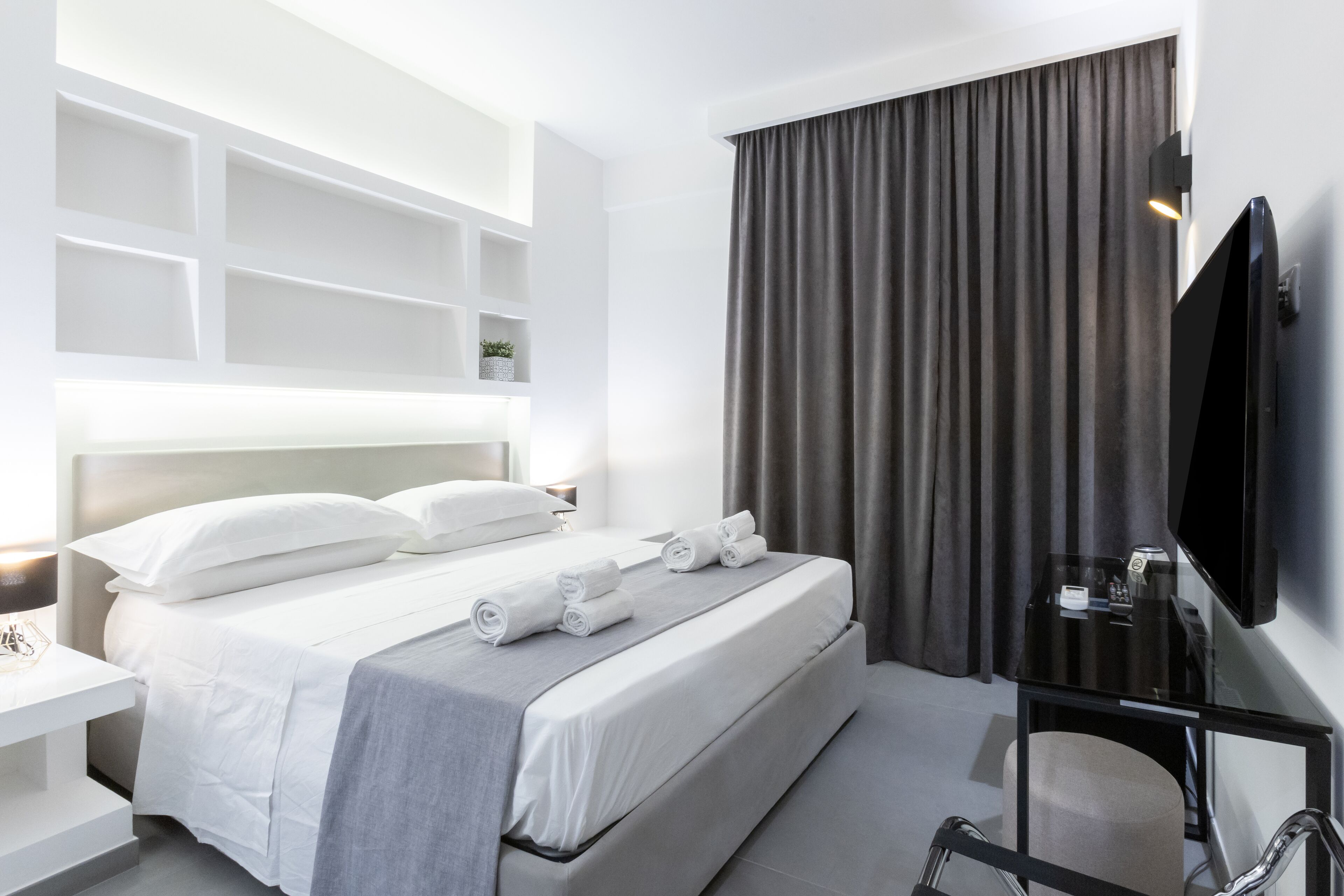 A modern hotel room with a neatly made bed, a flat-screen TV, a desk with a chair, and a large window with curtains.