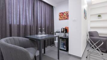 A modern living room with a dining table, two grey armchairs, a sofa, a coffee machine, and a small plant on a shelf.