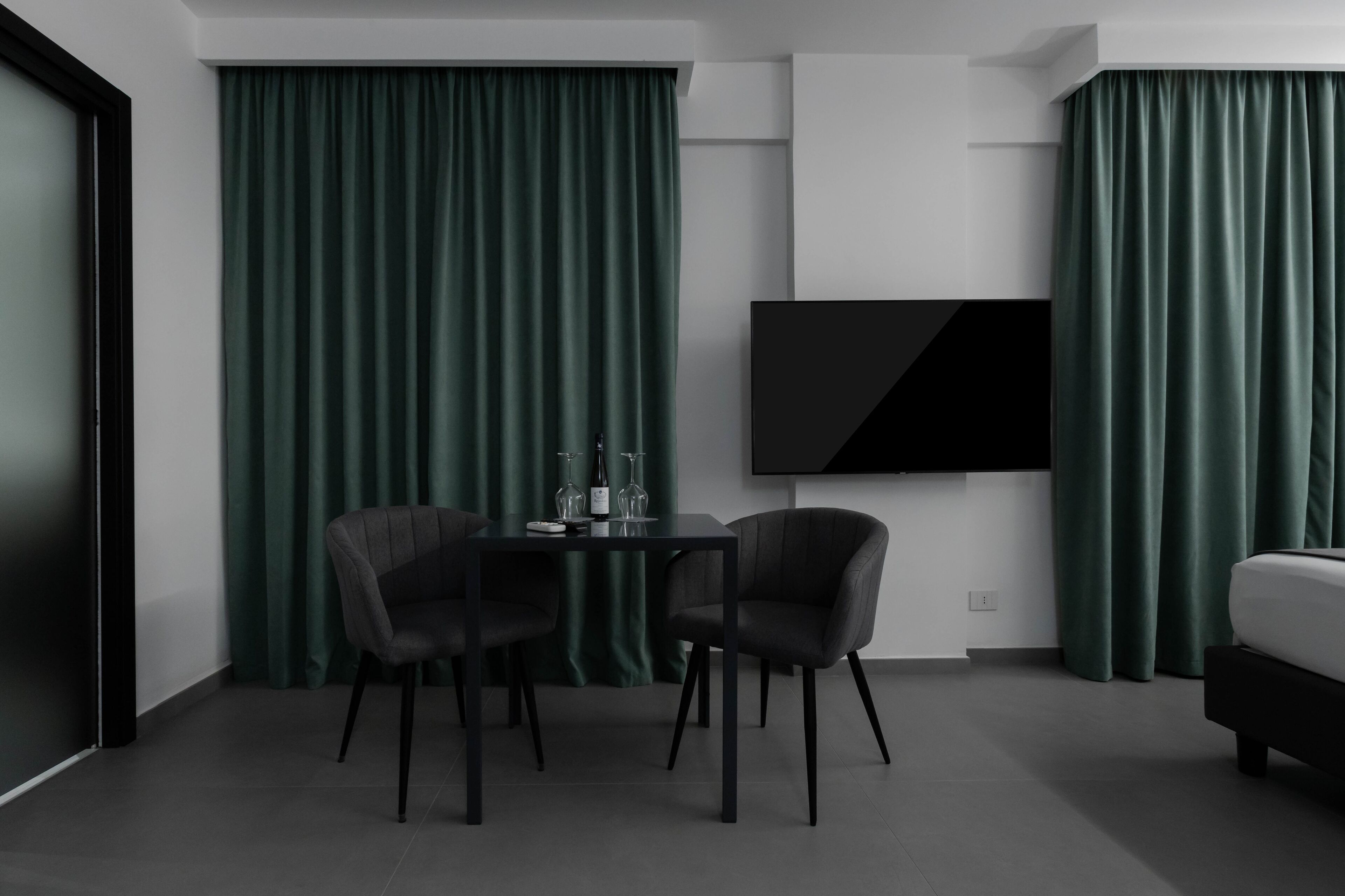 Una habitación de hotel moderna con un televisor de pantalla plana montado en la pared, una mesa de comedor con dos sillas y una cama con cabecero oscuro.