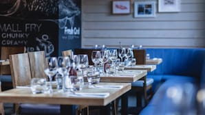 Interior de restaurante con mesas de madera listas para comer, un banco acolchado azul y un menú en pizarra al fondo.