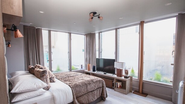 Una habitación de hotel moderna con una cama grande, un televisor de pantalla plana y vistas a la ciudad a través de amplios ventanales.