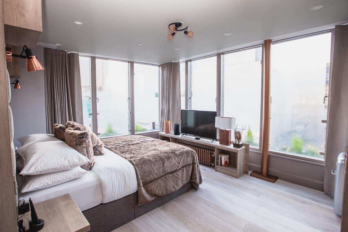 A modern hotel room with a large bed, a flat-screen TV, and a view of the city through large windows.
