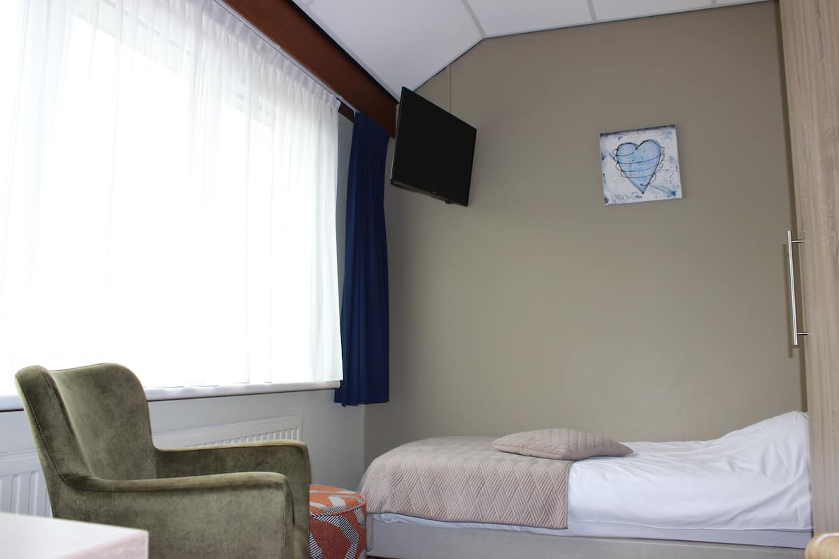 Una habitación con cama, una silla, televisor montado en la pared y ventana con cortinas.