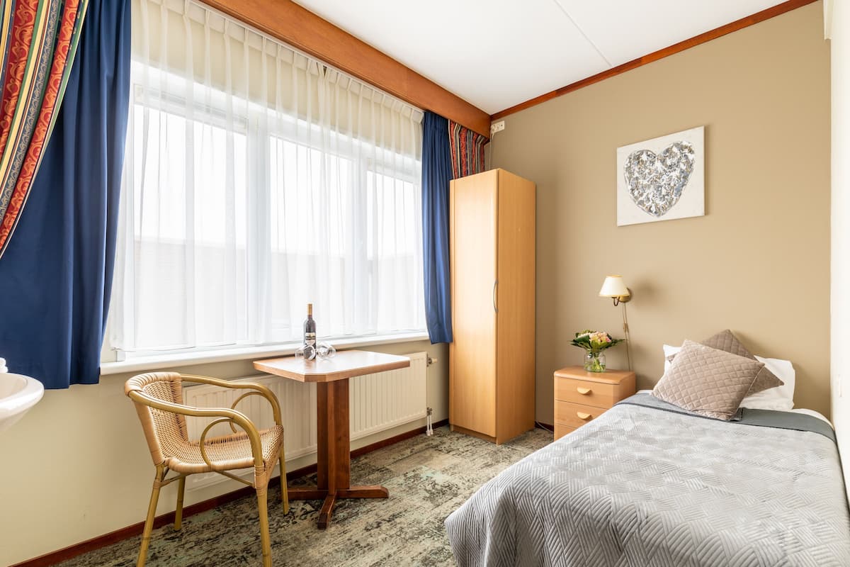 Habitación de hotel con cama, escritorio con silla, armario, mesita de noche con lámpara y ventana con cortinas.