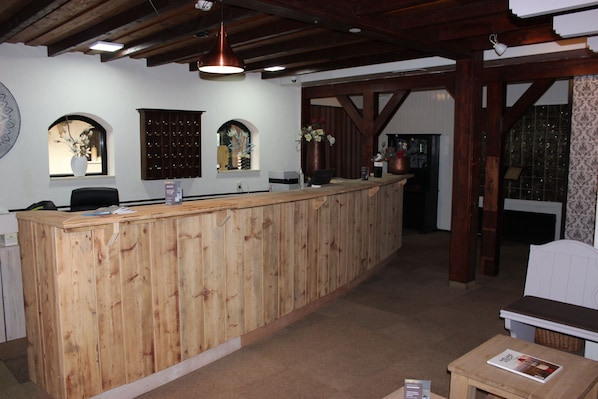 Un área de recepción con un mostrador de madera, una computadora y una silla. Hay jarrones decorativos en la pared y un papel tapiz estampado a la derecha.