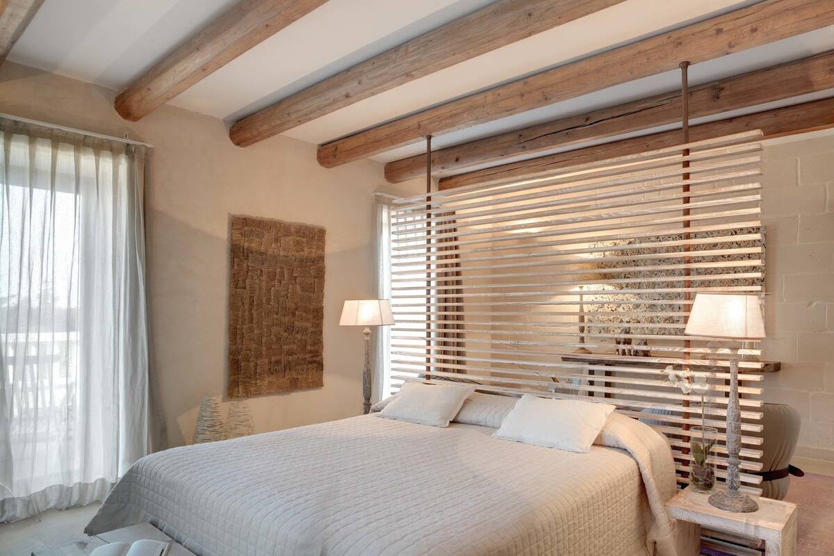 A bedroom with a large bed, wooden beams, a window with blinds, a bedside table with a lamp, and a wall-mounted artwork.