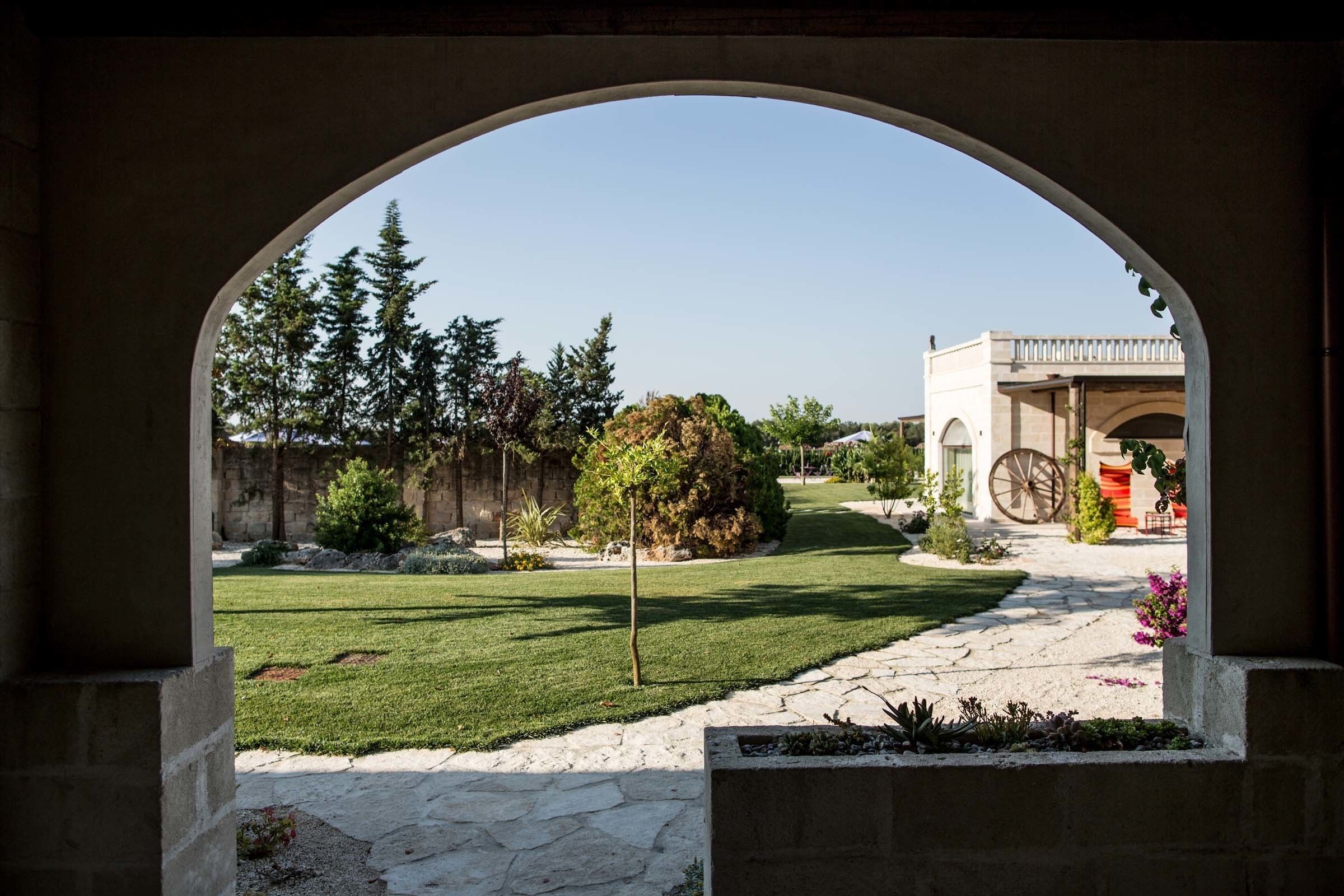 An arched entrance to a courtyard with a stone pathway, greenery, and a building with a large wheel in the background.