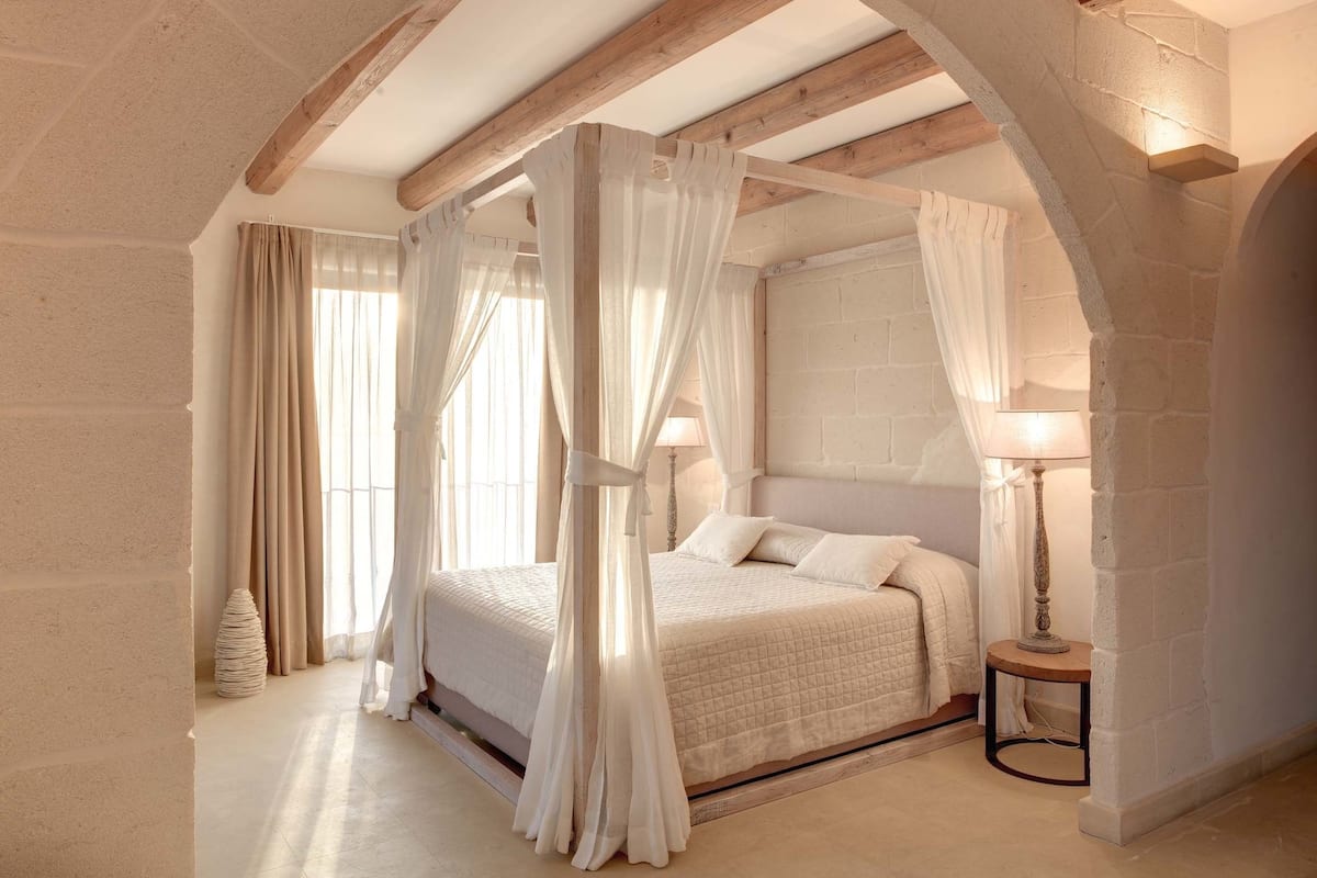 A bedroom with a four-poster bed, stone walls, and a wooden ceiling beam.
