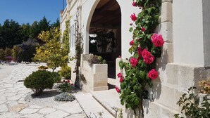 A building with a white facade, an arched entrance, and a garden with various plants and flowers. - Le Scuderie - Castello Monaci (Salice Salentino)