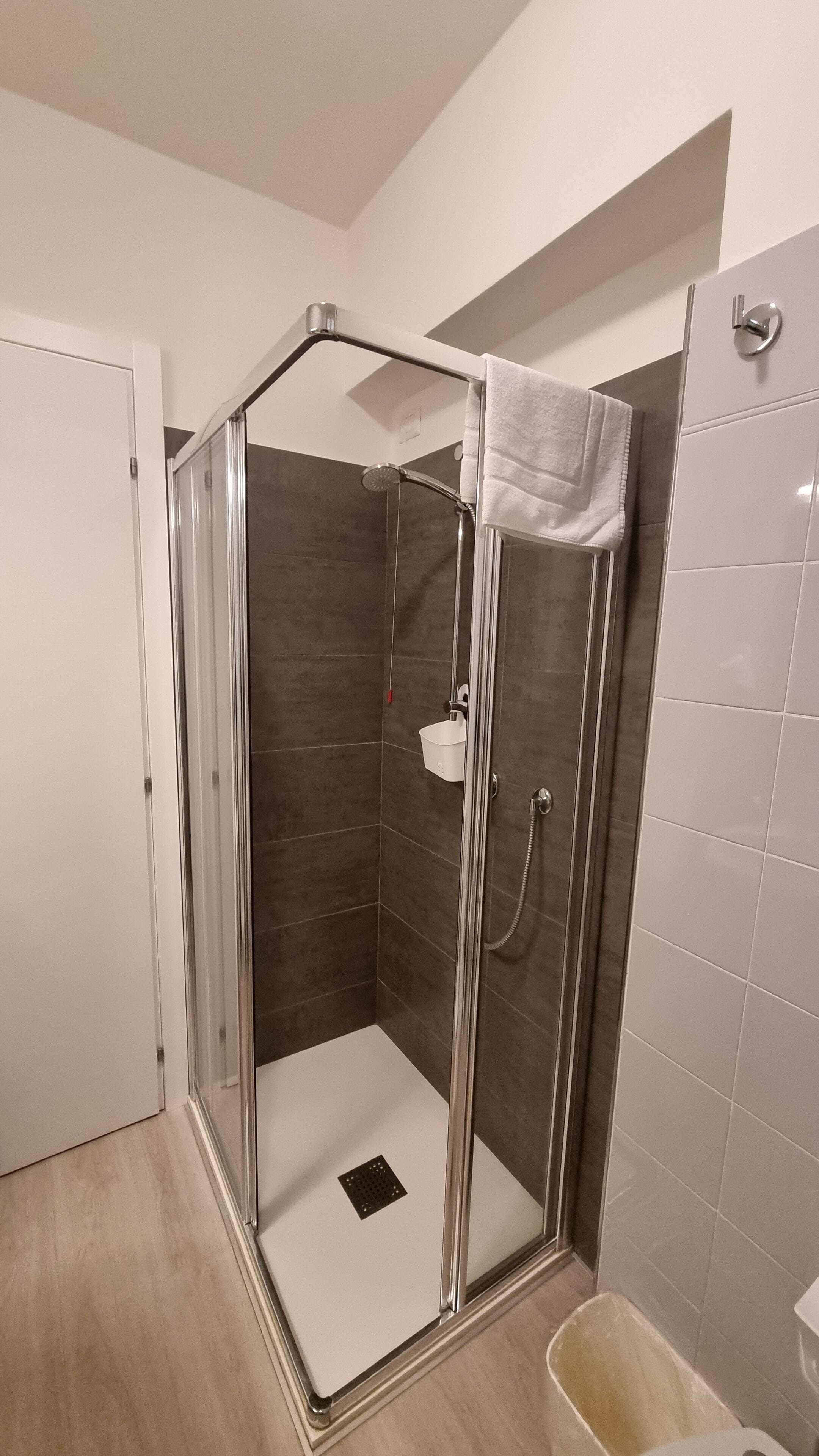 Une douche à l’italienne avec une porte en verre, une serviette blanche accrochée à la porte et un seau blanc fixé au mur.