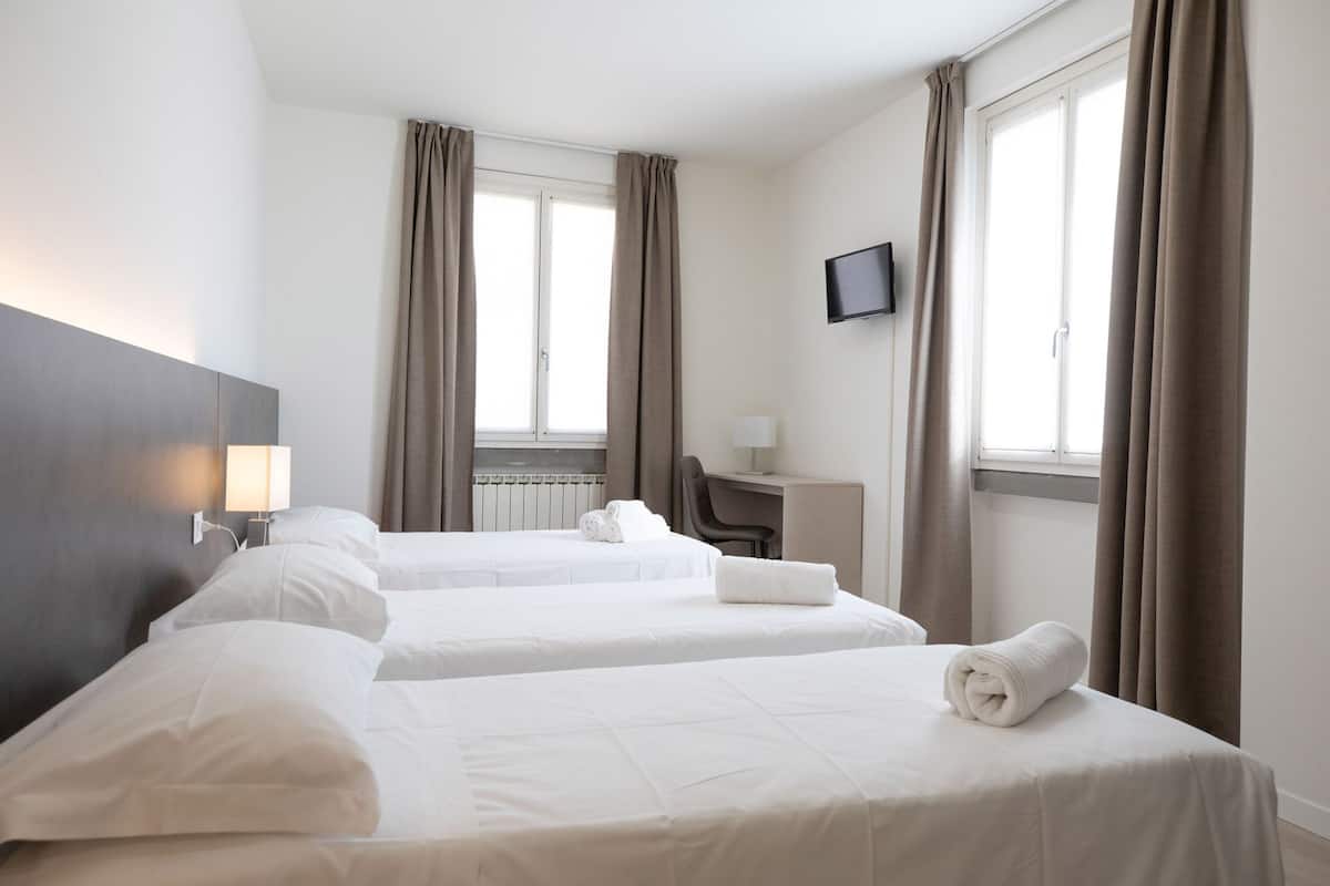 Habitación de hotel con dos camas, un escritorio, una silla, un televisor y ventanas grandes con cortinas.