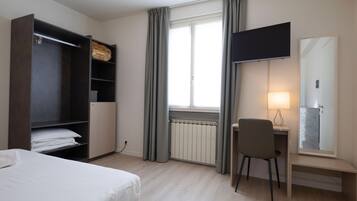 Une chambre d’hôtel comprenant un lit, un bureau avec une chaise, une télévision, une armoire et une fenêtre avec des rideaux.