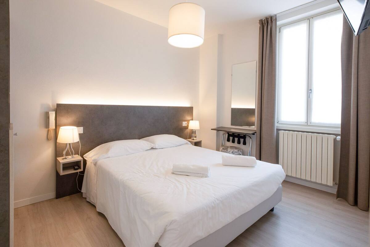 Habitación de hotel con una cama grande, dos lámparas de noche, una mesita, un espejo, una ventana con cortinas y un radiador.