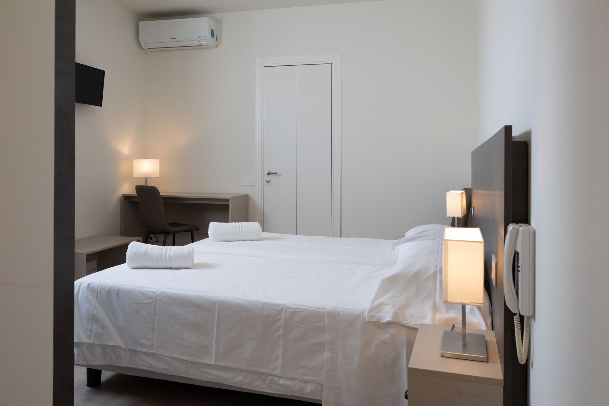 Ein Hotelzimmer mit Bett, Nachttischlampen, einem Schreibtisch mit Stuhl, einem Fernseher und einem Kleiderschrank.