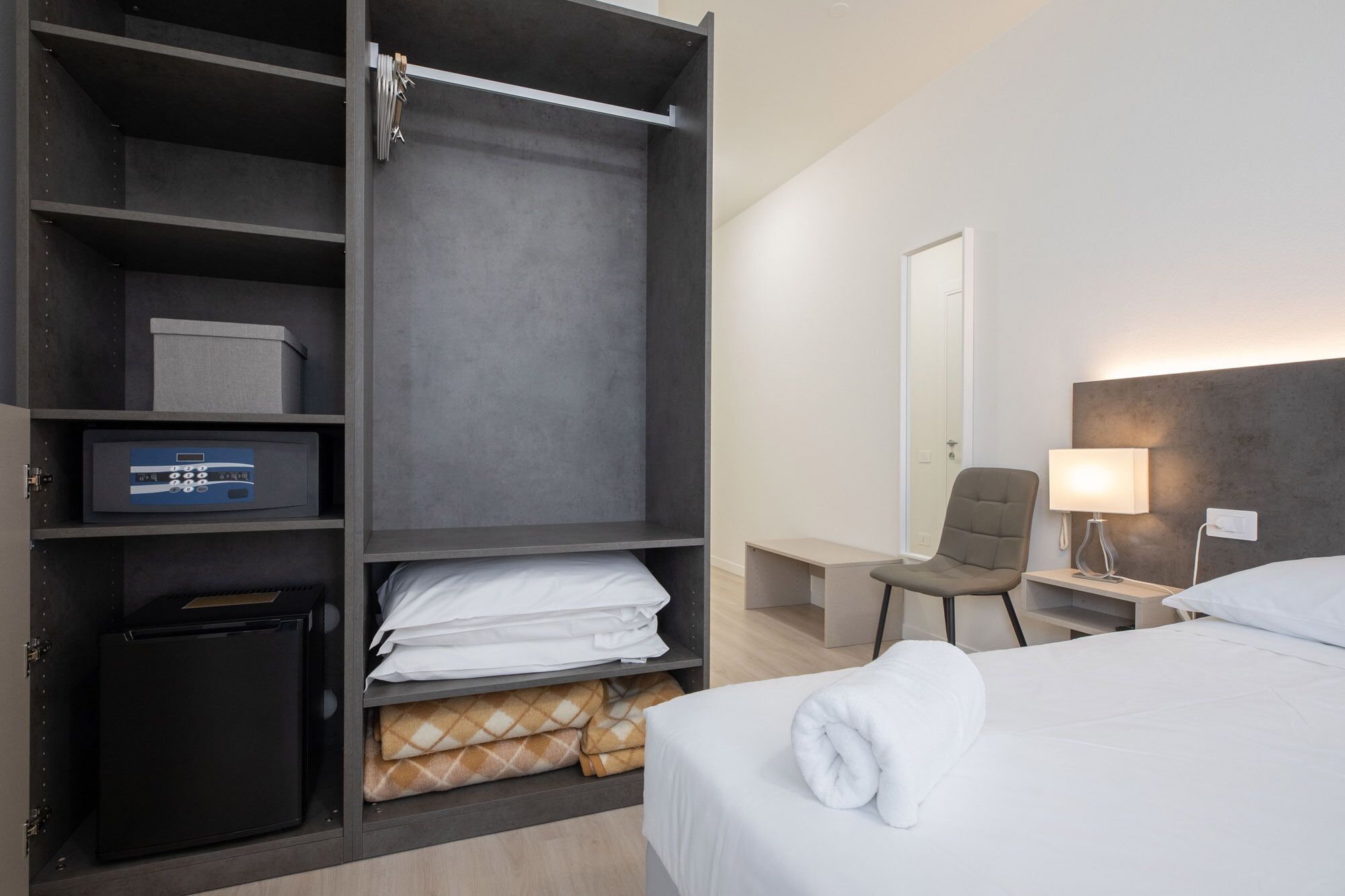 A hotel room with a bed, a desk with a lamp, a chair, a wardrobe with a safe, and a small table.