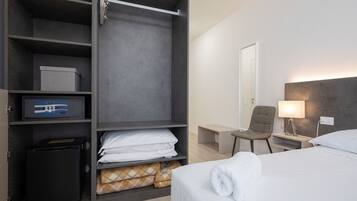 A hotel room with a bed, a desk with a lamp, a chair, a wardrobe with a safe, and a small table.