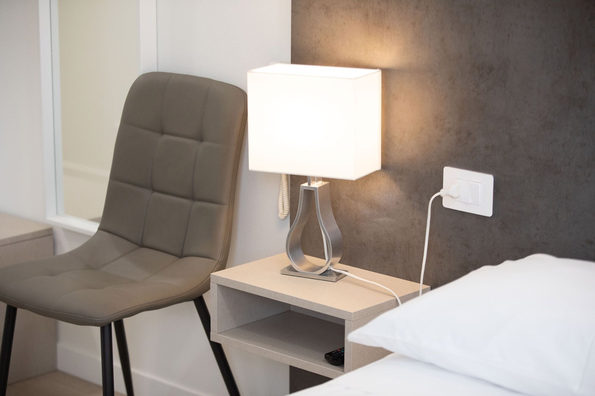 Ein modernes Hotelzimmer mit einem grau gepolsterten Sessel, einem Nachttisch mit Lampe und einem Bett mit weißer Bettwäsche.