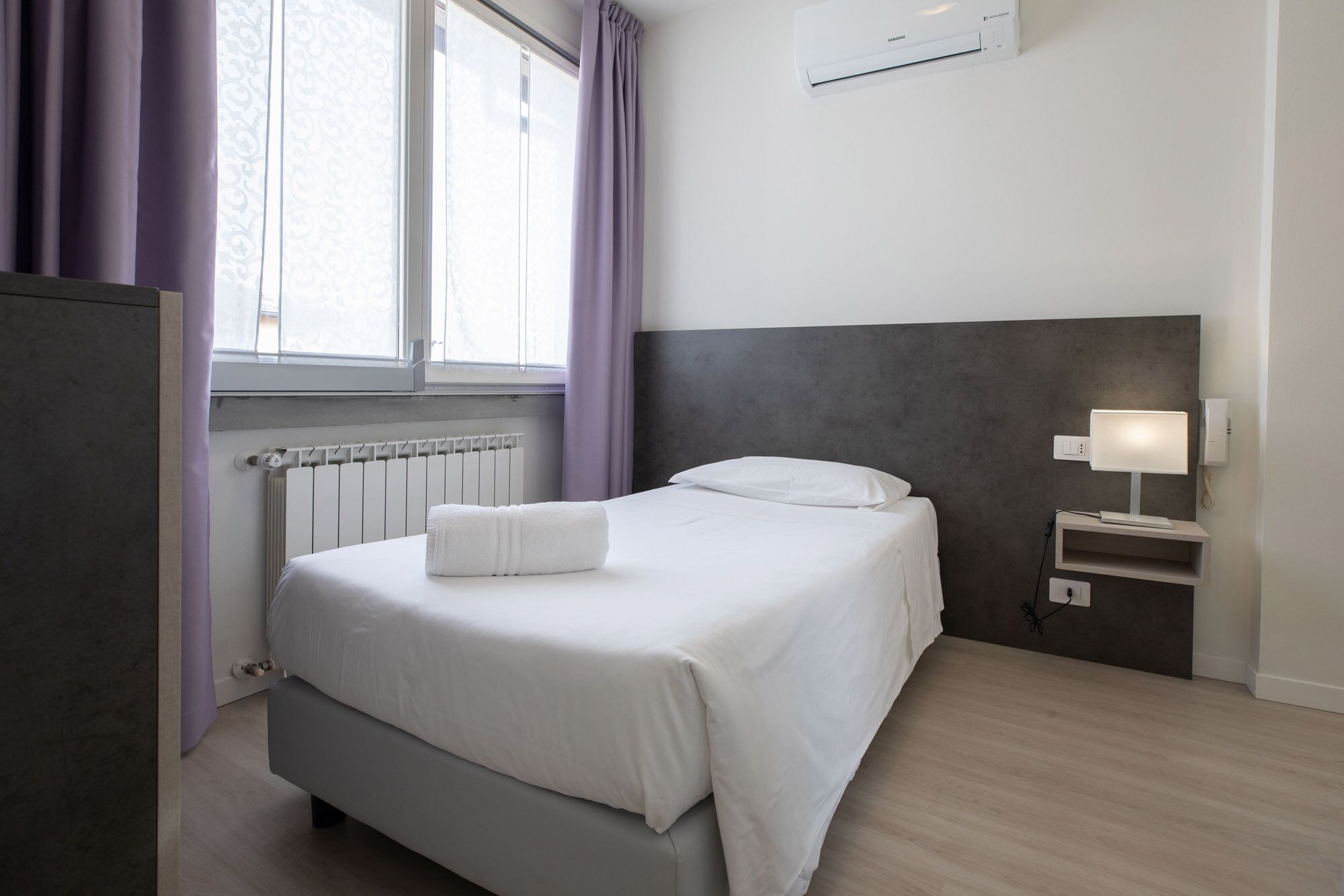 A neatly arranged hotel room with a bed, a bedside table with a lamp, a radiator, and an air conditioning unit.