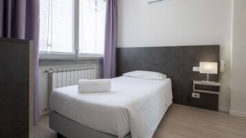 A neatly arranged hotel room with a bed, a bedside table with a lamp, a radiator, and an air conditioning unit.