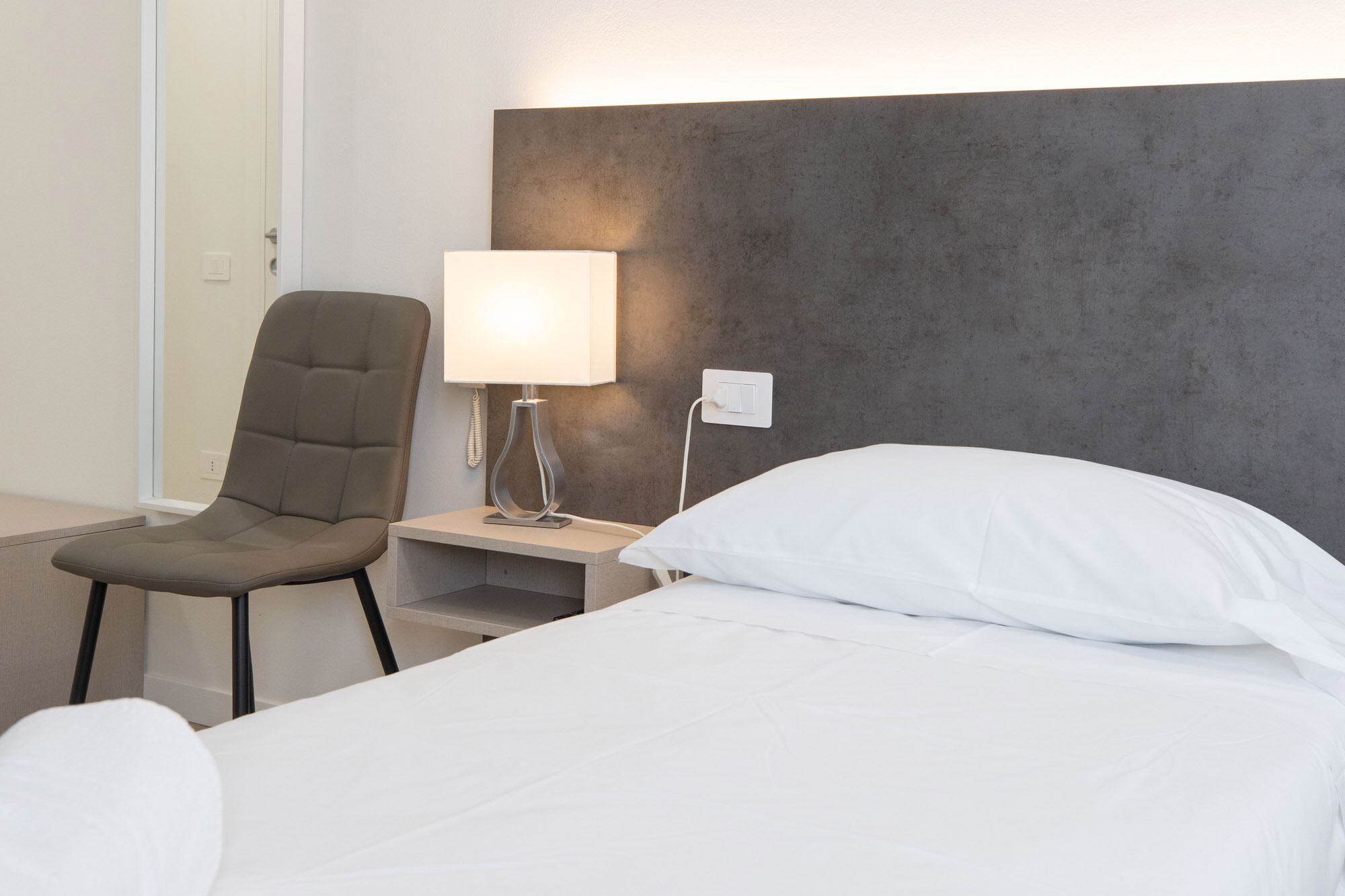 Ein Hotelzimmer mit Bett, Sessel, Nachttisch, Lampe und einer Steckdose.