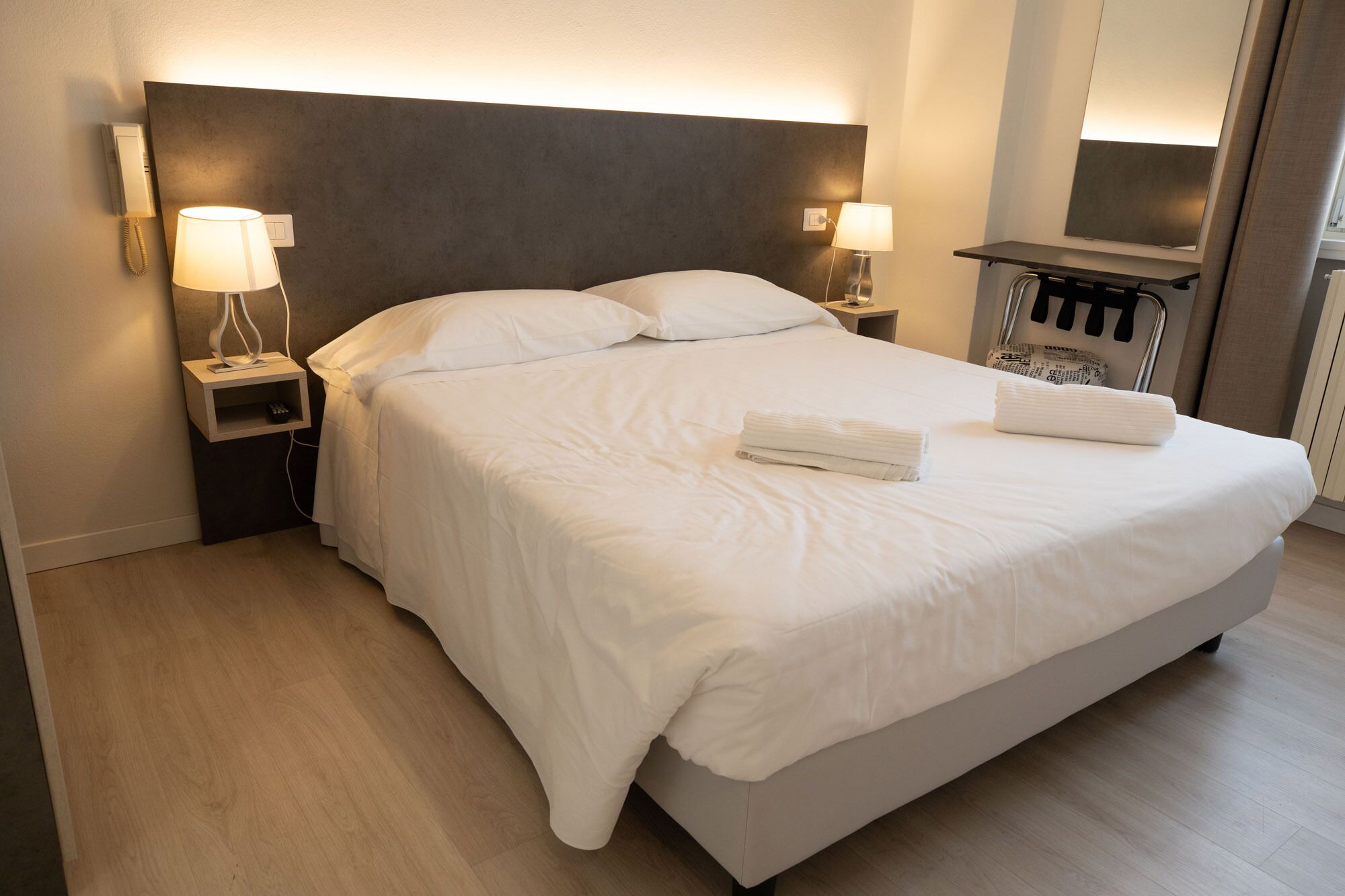A neatly made bed with white linens and two folded towels, a bedside table with a lamp, and a wall-mounted phone.