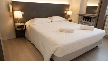 A neatly made bed with white linens and two folded towels, a bedside table with a lamp, and a wall-mounted phone.