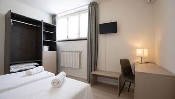 Ein Hotelzimmer mit Bett, Schreibtisch mit Stuhl, Fernseher, Kleiderschrank und Fenster mit Vorhängen.