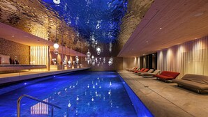A modern indoor pool with a blue-lit ceiling, seating area with red and beige chairs, and a reception desk. - Anewandter Historic Hotel (Gais)