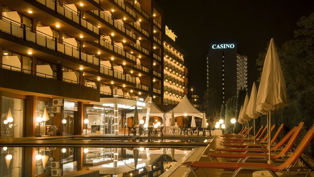 A hotel with a poolside area featuring lounge chairs and umbrellas, a casino sign in the background, and a modern building facade with balconies. - Gladiola Star (Golden Sands)