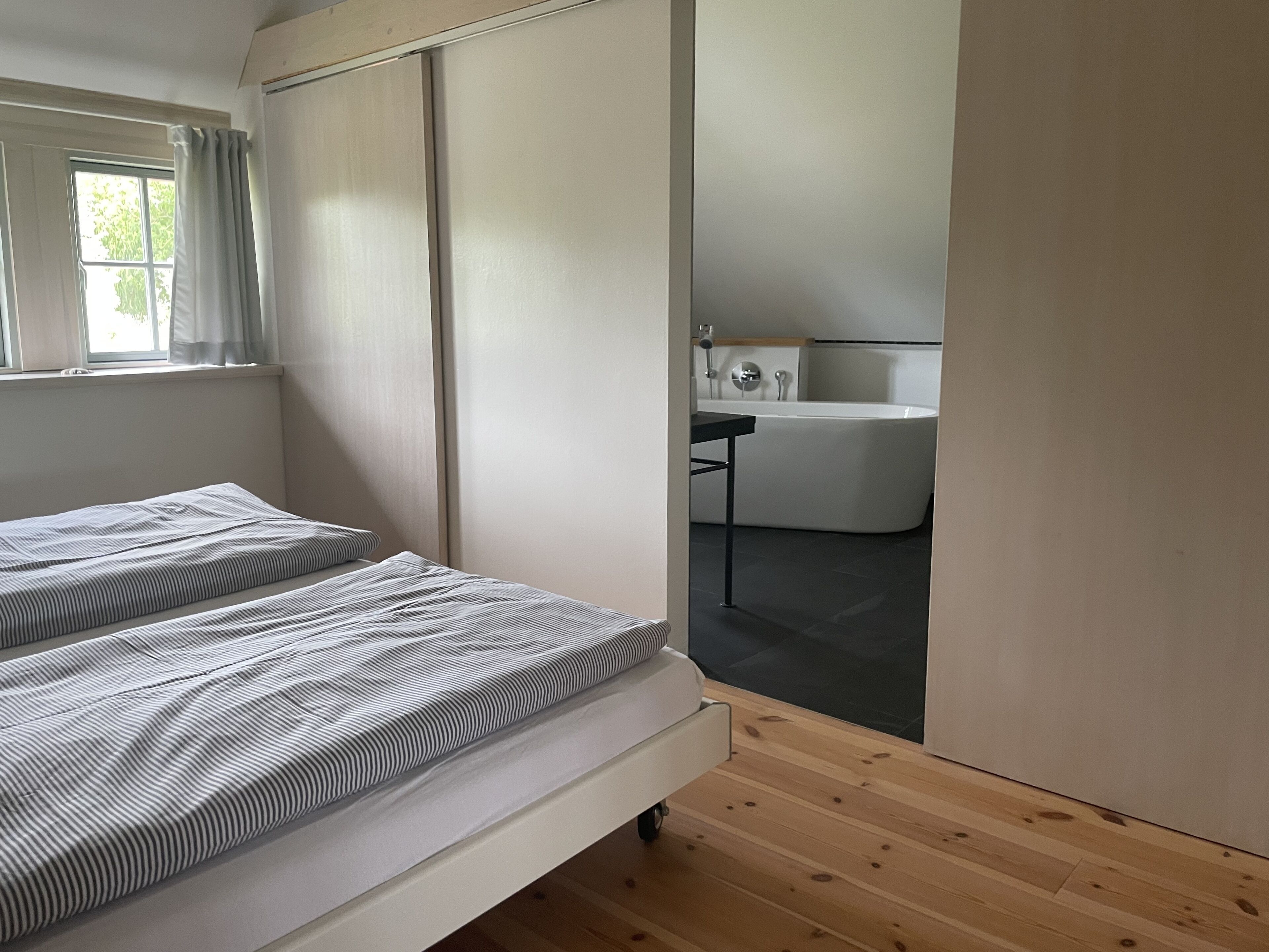 A bedroom with two beds, a window with curtains, a wooden floor, and a bathroom with a bathtub and sink.